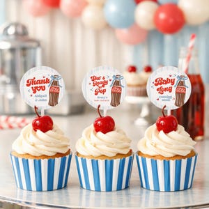 May include: Three cupcakes with white frosting, a cherry on top, and blue and white striped wrappers. Each cupcake has a circular topper with text and a vintage soda bottle graphic. Balloons in red, pink, and blue are in the background.