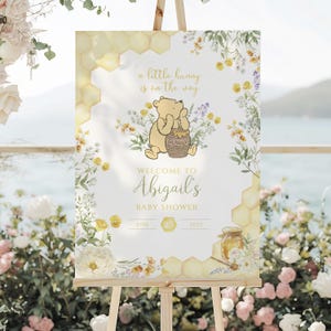 Puede incluir: Un letrero de bienvenida para baby shower sobre un caballete. El letrero presenta una ilustraci&oacute;n de Winnie the Pooh con el texto "a little hunny is on the way" y "Welcome to Abigail's Baby Shower". La fecha es el 25 de junio de 2025. Dise&ntilde;os florales y de panal enmarcan el texto.