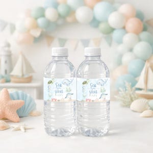 K&ouml;nnte beinhalten: Zwei transparente Plastikwasserflaschen mit wei&szlig;en Deckeln. Jede Flasche hat ein hellblaues Etikett mit Meeresbewohnern und dem Text "Sea you soon BABY". Der Hintergrund umfasst einen Ballonbogen und nautische Dekorationen.