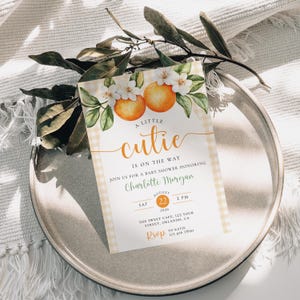 Peut inclure: Invitation de baby shower avec un motif floral orange et blanc. L'invitation pr&eacute;sente le texte "A Little Cutie is on the Way" et les d&eacute;tails de la baby shower de Charlotte Morgan. L'invitation est sur une assiette avec de la verdure.