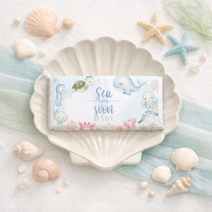 K&ouml;nnte beinhalten: Eine wei&szlig;e Schokoladentafel mit Babyparty-Motiv, mit Illustrationen von Meeresbewohnern und dem Text "Sea you soon BABY". Die Tafel wird auf einer muschelf&ouml;rmigen Schale pr&auml;sentiert, umgeben von Muscheln und Seesternen.
