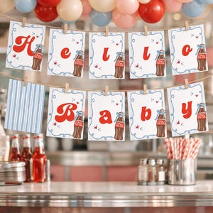 May include: A festive banner spelling "Hello Baby" with red letters and Coca-Cola bottle illustrations, strung across a retro diner setting. The banner is decorated with small hearts and is held by wooden clothespins. A colorful balloon arch adds to the celebratory atmosphere.