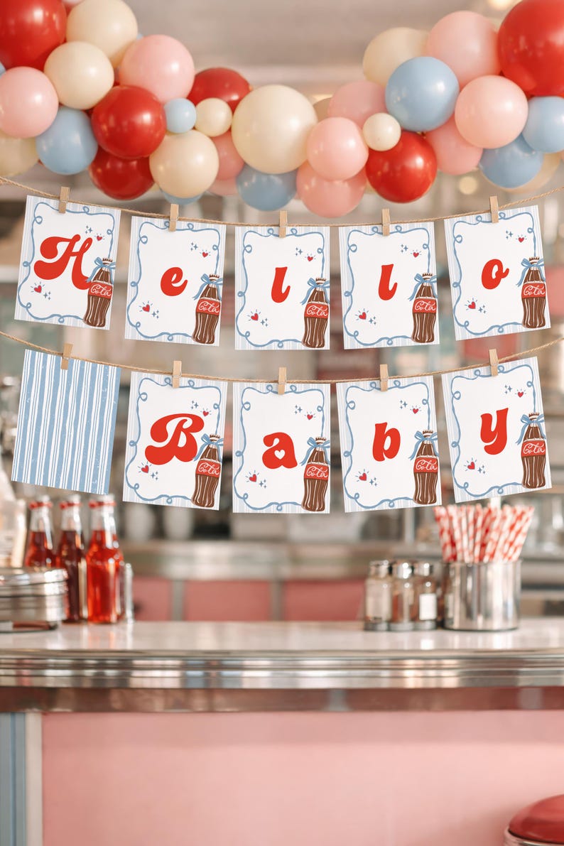 May include: A festive banner spelling "Hello Baby" with red letters and Coca-Cola bottle illustrations, strung across a retro diner setting. The banner is decorated with small hearts and is held by wooden clothespins. A colorful balloon arch adds to the celebratory atmosphere.