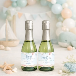 K&ouml;nnte beinhalten: Zwei gr&uuml;ne Champagnerflaschen mit silbernen Folienverschl&uuml;ssen, jede mit einem individuellen Etikett. Auf einer steht "Thank you" und auf der anderen "Jennifer's Baby Shower". Der Hintergrund umfasst einen Ballonbogen, ein Spielzeugsegelschiff und Muscheln.