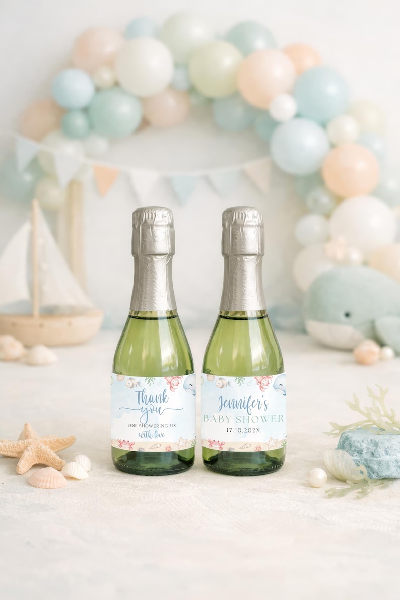 K&ouml;nnte beinhalten: Zwei gr&uuml;ne Champagnerflaschen mit silbernen Folienverschl&uuml;ssen, jede mit einem individuellen Etikett. Auf einer steht "Thank you" und auf der anderen "Jennifer's Baby Shower". Der Hintergrund umfasst einen Ballonbogen, ein Spielzeugsegelschiff und Muscheln.