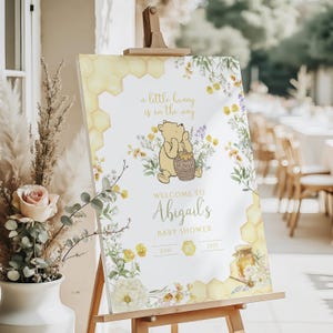 Puede incluir: Un letrero de bienvenida para baby shower con Winnie the Pooh. El letrero incluye el texto "a little hunny is on the way" y "Welcome to Abigail's Baby Shower". Decorado con dise&ntilde;os florales y de panal.