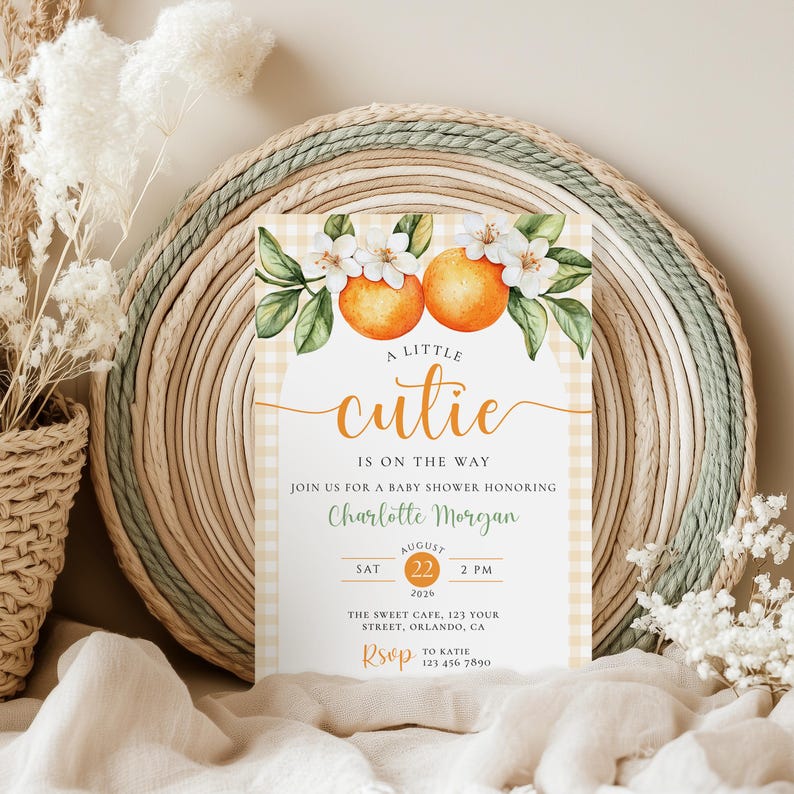 Peut inclure: Faire-part de baby shower avec un motif floral orange et blanc. L'invitation pr&eacute;sente le texte "A Little Cutie Is On The Way" et les d&eacute;tails de l'&eacute;v&eacute;nement. L'invitation est plac&eacute;e sur un fond tiss&eacute; avec un motif vichy.