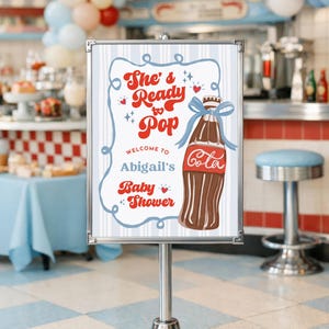 May include: A sign for Abigail's baby shower with a retro design. The sign features the text "She's Ready to Pop" and a cartoon illustration of a cola bottle tied with a blue ribbon. The background includes a diner setting with balloons.