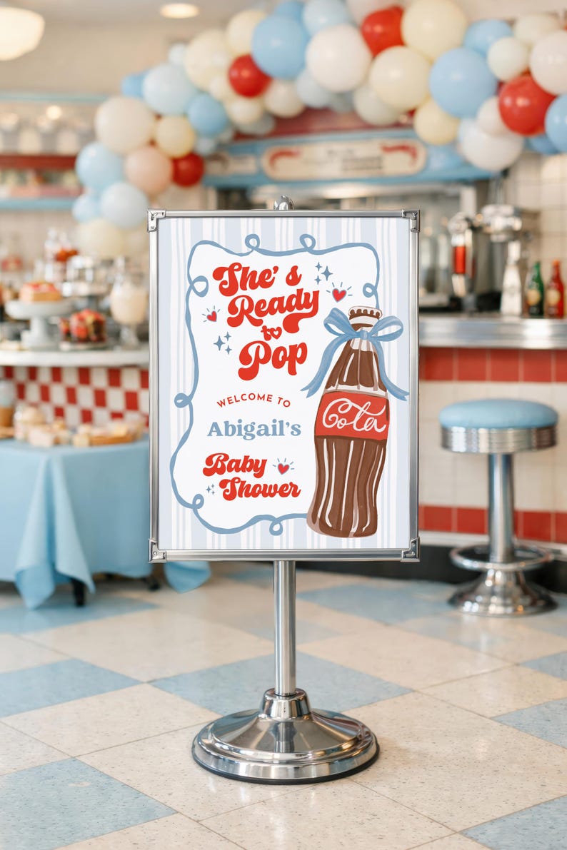 May include: A sign for Abigail's baby shower with a retro design. The sign features the text "She's Ready to Pop" and a cartoon illustration of a cola bottle tied with a blue ribbon. The background includes a diner setting with balloons.