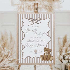 Op de afbeelding: Een welkomstbord voor een babyshower op een houten ezel. Het bord heeft een gestreepte achtergrond en een decoratieve rand. De tekst luidt "We Can Bearly Wait, Welcome to Anastasia's Baby Shower" met een illustratie van een teddybeer. De datum is 22 november 2026 om 14:00 uur.