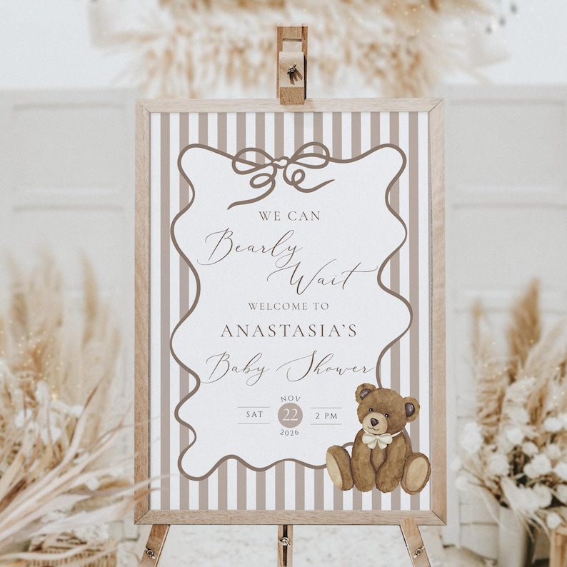 Op de afbeelding: Een welkomstbord voor een babyshower op een houten ezel. Het bord heeft een gestreepte achtergrond en een decoratieve rand. De tekst luidt "We Can Bearly Wait, Welcome to Anastasia's Baby Shower" met een illustratie van een teddybeer. De datum is 22 november 2026 om 14:00 uur.
