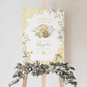 Puede incluir: Un letrero de bienvenida para baby shower con Winnie the Pooh y el texto "a little hunny is on the way." El letrero incluye dise&ntilde;os florales y de panal, con "Abigail's" y la fecha 25 de junio de 2025.