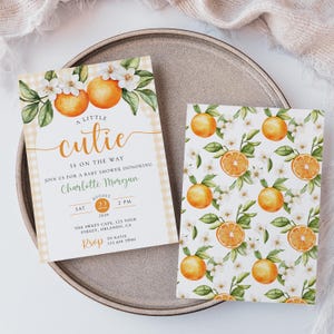 Peut inclure: Invitations de baby shower avec des oranges &agrave; l'aquarelle, des fleurs et des feuilles vertes. L'invitation principale indique "A Little Cutie is on the Way" avec les d&eacute;tails de l'&eacute;v&eacute;nement. Une carte assortie affiche un motif d'orange et de fleurs. Les invitations sont sur un plateau gris.