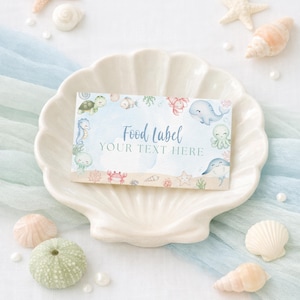 K&ouml;nnte beinhalten: Ein rechteckiges Lebensmitteletikett mit einem Aquarelldesign von Meeresbewohnern, darunter eine Schildkr&ouml;te, ein Seepferdchen und ein Wal. Das Etikett tr&auml;gt die Aufschrift "Food Label" und "Your Text Here" in eleganter Schrift. Das Etikett liegt auf einem wei&szlig;en, muschelf&ouml;rmigen Teller.