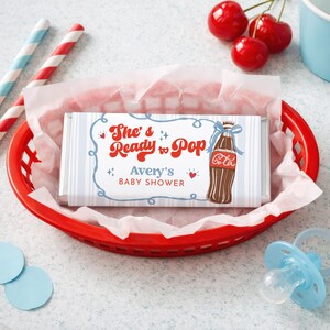 May include: A red plastic basket holds a candy bar wrapped in a blue and white striped wrapper. The wrapper reads "She's Ready to Pop" with a cola bottle graphic, for a baby shower. Red and white straws, cherries, and a blue pacifier are also in the image.