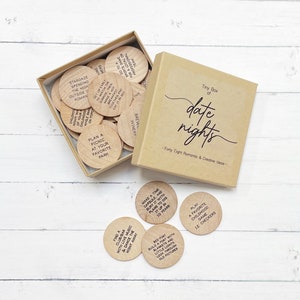 May include: A brown box with the text "Tiny Box of date nights - Forty Eight Romantic & Creative Ideas" on the front. The box contains a set of wooden tokens with different date night ideas written on them.