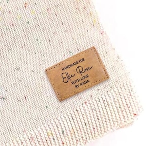 May include: Close-up of a light-colored knit beanie with flecks of color. A rectangular brown leather tag is sewn on, with the words "HANDMADE FOR Ellie Rose WITH LOVE BY NANA" in black script.