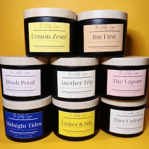 Può includere: Una collezione di sette candele nere in miscela di cera di soia con coperchi in legno. Ogni candela ha un'etichetta con un nome e una descrizione della fragranza unici, come "Lemon Zesté" e "But First". Le etichette sono in vari colori, tra cui giallo, rosa e blu.