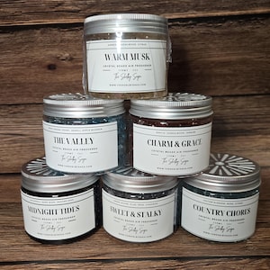 May include: A stack of six glass jars with silver lids, each containing crystal beads air fresheners. The jars have white labels with black text, including the scent names: Warm Musk, The Valley, Charm & Grace, Midnight Tides, Sweet & Stalky, and Country Chores.