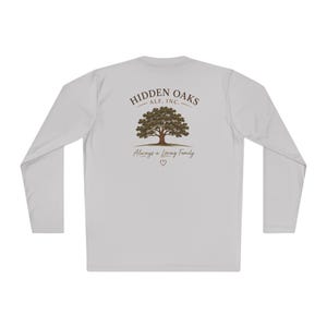 May include: A light gray long-sleeve shirt featuring a graphic on the back. The design includes a brown oak tree with green leaves, the text "HIDDEN OAKS ALF, INC." above it, and the phrase "Always a Loving Family" with a small heart below.