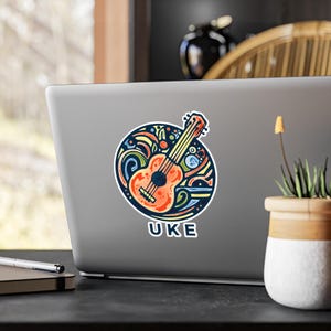 May include: A colorful sticker featuring a ukulele illustration. The ukulele is orange with black details, surrounded by a blue and yellow design. The word "UKE" is printed in white on a blue background.