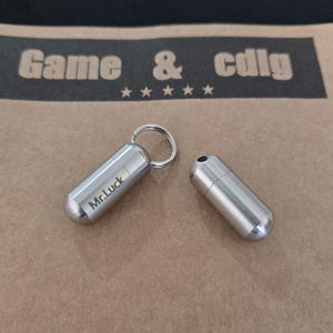 Puede incluir: Dos pequeños pastilleros metálicos plateados. Uno está unido a un llavero y tiene grabado "Mr.Luck". El otro recipiente está abierto, revelando un interior oscuro. El fondo presenta el texto "Game & cdlg" sobre cinco estrellas.