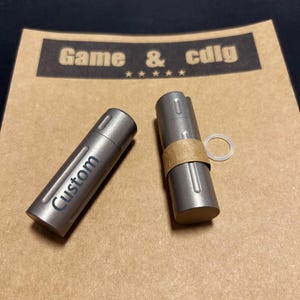 Könnte beinhalten: Zwei zylindrische, metallisch graue Behälter mit mattem Finish. Einer trägt die Gravur "Custom". Der andere hat ein braunes Papierband und einen transparenten, kreisförmigen Ring. Der Hintergrund ist ein braunes Papier mit dem Text "Game & cdig".