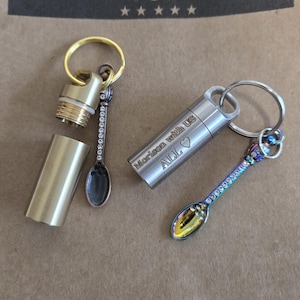 Puede incluir: Dos llaveros metálicos con pequeñas cucharas. Uno es dorado, el otro plateado. El llavero dorado está desenroscado, revelando un compartimento. El llavero plateado tiene la inscripción "Morison with us ALL ❤".