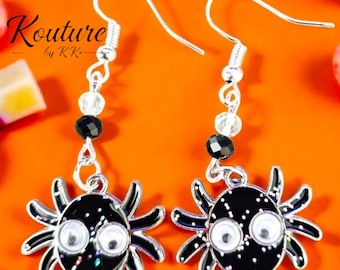 May include: A pair of silver-toned hook earrings featuring black spider charms with googly eyes and glitter accents. Each earring includes a clear bead and a black faceted bead on the wire. The text Kouture by KK is visible in the upper left corner.