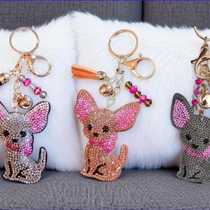 May include: Three rhinestone Chihuahua keychains. Each dog has pink-lined ears and a pink collar. The keychains have gold-colored hardware, tassels, and decorative beads. One dog is silver, one is brown, and one is dark gray.