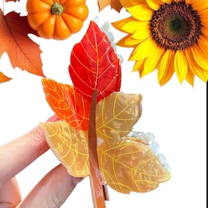 Peut inclure: Une pince à cheveux sur le thème de l'automne avec des motifs de feuilles aux tons rouges, oranges et dorés. La pince a une tige marron et un fond blanc. Une citrouille et un tournesol sont en arrière-plan.