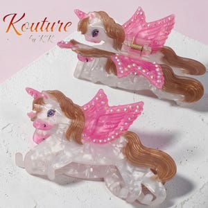 Peut inclure: Deux pinces &agrave; cheveux en forme de licorne au design marbr&eacute; blanc avec des crini&egrave;res brunes, des ailes roses et des cristaux iris&eacute;s. Le texte Kouture by KK est visible dans le coin sup&eacute;rieur gauche. Chaque pince poss&egrave;de un ressort m&eacute;tallique.
