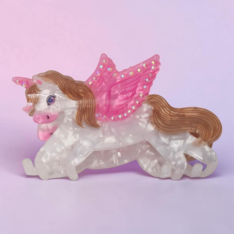Peut inclure: Une pince &agrave; cheveux en forme de licorne ail&eacute;e avec un corps blanc nacr&eacute;, une crini&egrave;re et une queue brunes, et des ailes roses translucides. Les ailes pr&eacute;sentent de petites paillettes iris&eacute;es incrust&eacute;es, et la licorne a un museau et une corne roses.