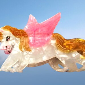 Peut inclure: Une pince &agrave; cheveux en forme de licorne ail&eacute;e avec un corps blanc nacr&eacute;, une crini&egrave;re et une queue brun dor&eacute;, et des ailes roses translucides. La licorne pr&eacute;sente une corne unique sur le front et un visage d&eacute;taill&eacute; avec des yeux sombres et un nez rose.