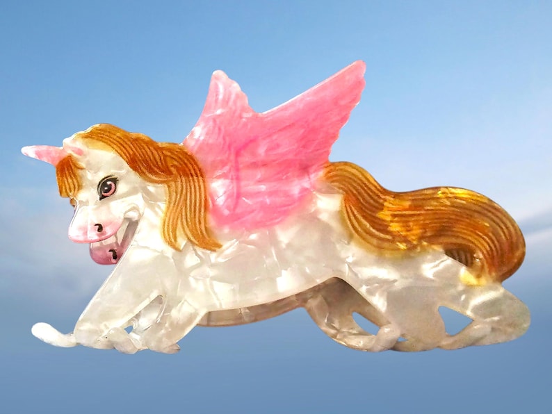 Peut inclure: Une pince &agrave; cheveux en forme de licorne ail&eacute;e avec un corps blanc nacr&eacute;, une crini&egrave;re et une queue brun dor&eacute;, et des ailes roses translucides. La licorne pr&eacute;sente une corne unique sur le front et un visage d&eacute;taill&eacute; avec des yeux sombres et un nez rose.