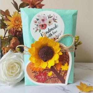 Peut inclure: Un paquet en papier turquoise clair orn&eacute; d'un autocollant rond avec des illustrations florales et le texte Handmade with love. Un tournesol en papier jaune et une carte en forme de c&oelig;ur avec des feuilles d'automne sont li&eacute;s par une ficelle.