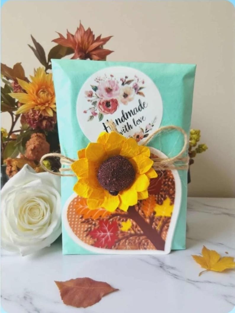 Peut inclure: Un paquet en papier turquoise clair orn&eacute; d'un autocollant rond avec des illustrations florales et le texte Handmade with love. Un tournesol en papier jaune et une carte en forme de c&oelig;ur avec des feuilles d'automne sont li&eacute;s par une ficelle.