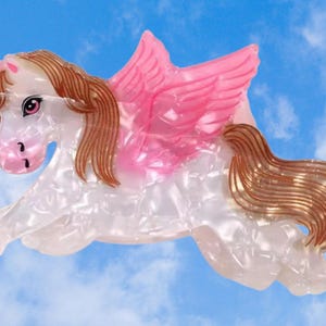 Peut inclure: Une pince &agrave; cheveux en forme de licorne ail&eacute;e avec un corps blanc nacr&eacute;, une corne rose clair, des ailes roses translucides, ainsi qu'une crini&egrave;re et une queue brunes. La licorne est repr&eacute;sent&eacute;e au galop avec des d&eacute;tails faciaux et des yeux roses.