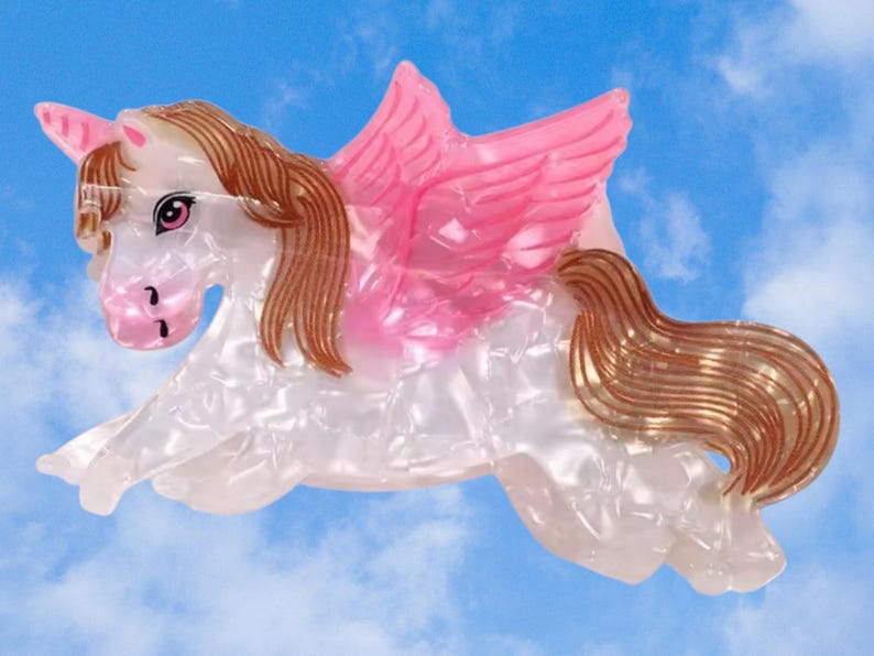 Peut inclure: Une pince &agrave; cheveux en forme de licorne ail&eacute;e avec un corps blanc nacr&eacute;, une corne rose clair, des ailes roses translucides, ainsi qu'une crini&egrave;re et une queue brunes. La licorne est repr&eacute;sent&eacute;e au galop avec des d&eacute;tails faciaux et des yeux roses.
