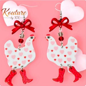 May include: A pair of dangle earrings featuring white chicken charms with pink and red heart patterns. Each chicken wears red glittery boots. Above the chicken, a red bow, a clear bead, and a red bead connect to a silver hook. The text Kouture by KK is visible.