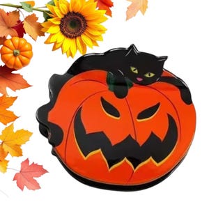 以下が含まれることがあります： ハロウィーンをテーマにした装飾で、笑顔のオレンジ色のカボチャと、その上に休んでいる黒猫が特徴です。猫は黄色い目をしています。背景にはヒマワリ、小さなカボチャ、秋の葉が含まれています。