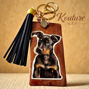 Puede incluir: Un llavero rectangular de resina marrón con una ilustración de un cachorro negro y fuego, una borla de piel sintética negra y una anilla de metal dorado con un pequeño dije en forma de corazón. El texto Kouture by KK es visible arriba a la derecha.