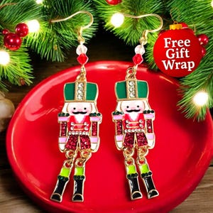 May include: A pair of gold-toned dangle earrings featuring a festive nutcracker design. The earrings have a red, green, pink, and white color scheme with a sparkling, glittery appearance. The earrings are displayed on a red plate with Christmas tree branches in the background.