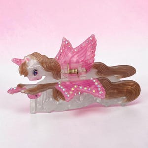 Peut inclure: Une pince &agrave; cheveux en forme de licorne ail&eacute;e blanche translucide avec une crini&egrave;re et une queue brunes. Les ailes sont orn&eacute;es de d&eacute;tails roses et de petits strass iris&eacute;s. Un m&eacute;canisme &agrave; ressort en m&eacute;tal dor&eacute; est visible au centre.