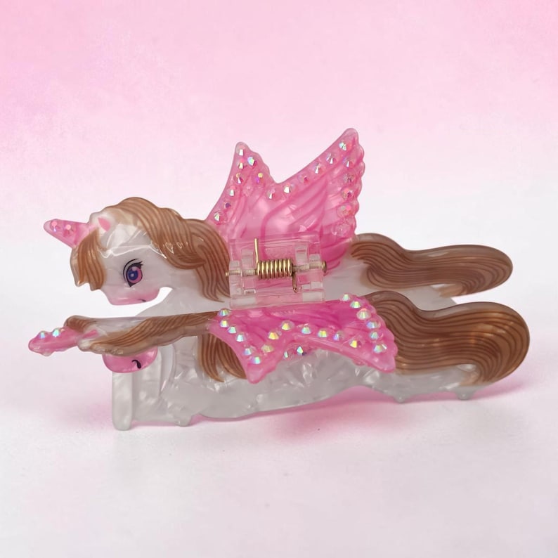 Peut inclure: Une pince &agrave; cheveux en forme de licorne ail&eacute;e blanche translucide avec une crini&egrave;re et une queue brunes. Les ailes sont orn&eacute;es de d&eacute;tails roses et de petits strass iris&eacute;s. Un m&eacute;canisme &agrave; ressort en m&eacute;tal dor&eacute; est visible au centre.