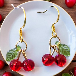 Puede incluir: Pendientes dorados con cuentas rojas en forma de cereza y detalles de hojas verdes. Los pendientes se exhiben en un plato blanco, rodeados de cerezas frescas. La longitud total es de aproximadamente 3,8 cm.