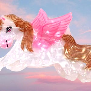 Peut inclure: Une pince &agrave; cheveux en forme de licorne ail&eacute;e. Le corps est en mat&eacute;riau blanc nacr&eacute; avec une corne rose, des ailes roses, ainsi qu'une crini&egrave;re et une queue brunes. La licorne est repr&eacute;sent&eacute;e dans une pose bondissante avec des traits d&eacute;taill&eacute;s.