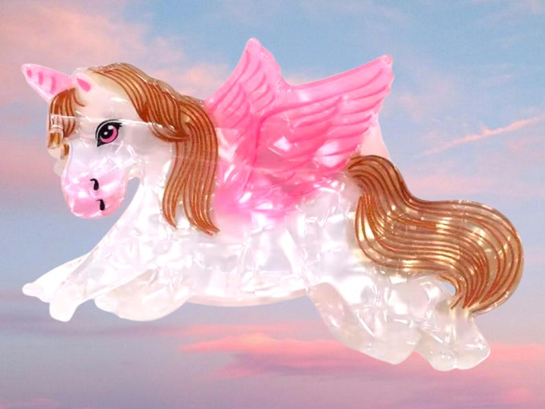 Peut inclure: Une pince &agrave; cheveux en forme de licorne ail&eacute;e. Le corps est en mat&eacute;riau blanc nacr&eacute; avec une corne rose, des ailes roses, ainsi qu'une crini&egrave;re et une queue brunes. La licorne est repr&eacute;sent&eacute;e dans une pose bondissante avec des traits d&eacute;taill&eacute;s.