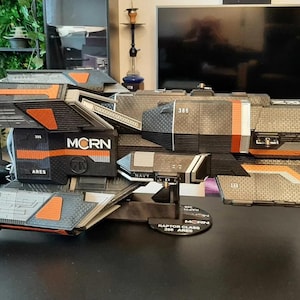 以下が含まれることがあります： 側面に「MCRN」と「ARES」の文字が書かれた黒とオレンジの宇宙船の模型。模型は、「RAPTOR CLASS - ARES」と書かれた白いラベルが付いた黒いスタンドの上に置かれている