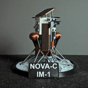 May include: A 3D printed model of the Nova-C rocket, a proposed lunar lander. The model is on a circular base with a white surface that resembles the moon. The base has a black label with the text "NOVA-C IM-1".